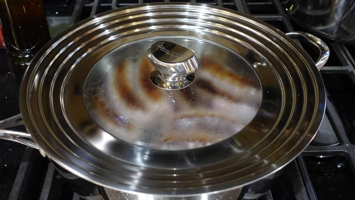 A covered skillet on the stovetop filled with pan seared bratwurst.