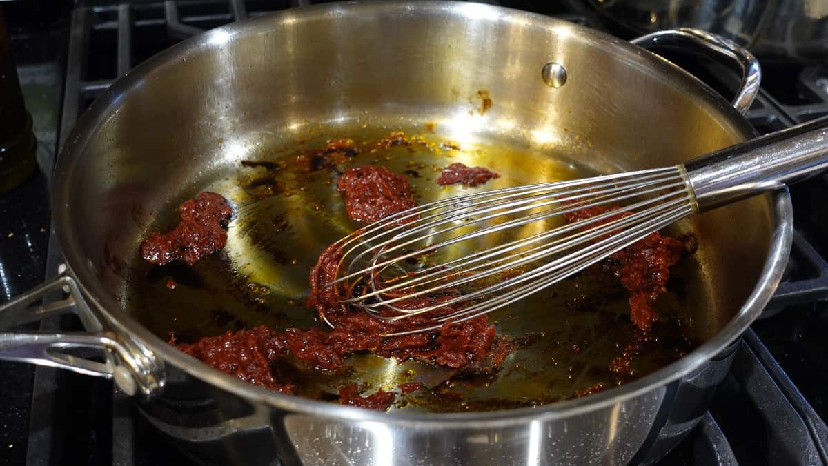 Tomato past being added to a skillet with a whisk.