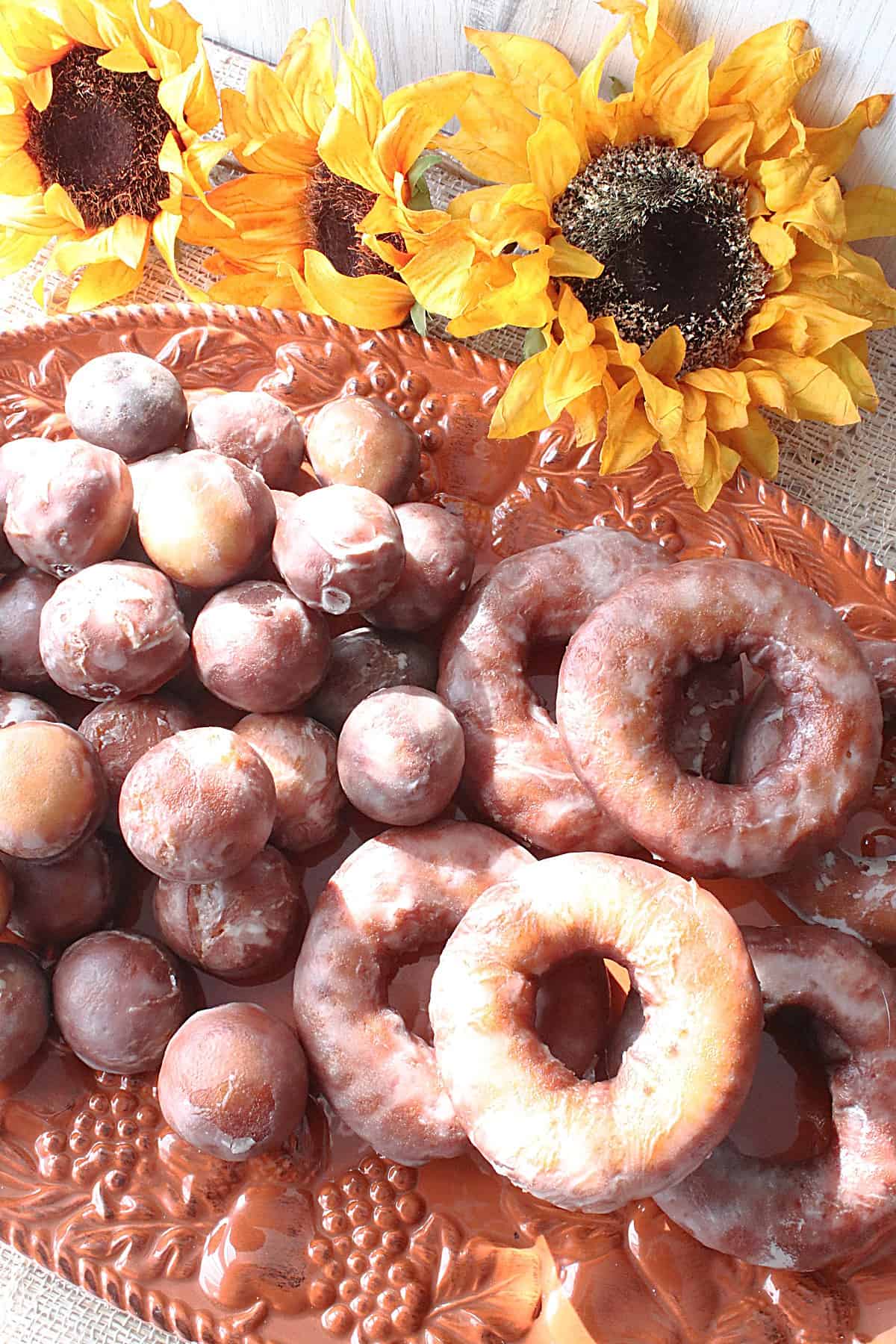 Donut holes and Glazed Apple Cider Donuts on an orange platter with sunflowers in the background.