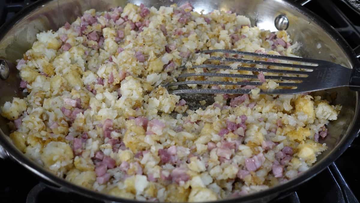 Broken up tater tots and diced ham in a skillet with a spatula.
