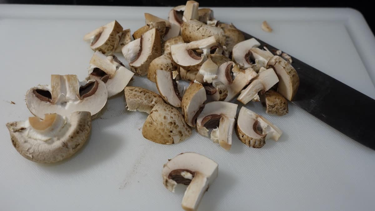 Sliced and quartered baby bella mushrooms on a cutting board with a knife.