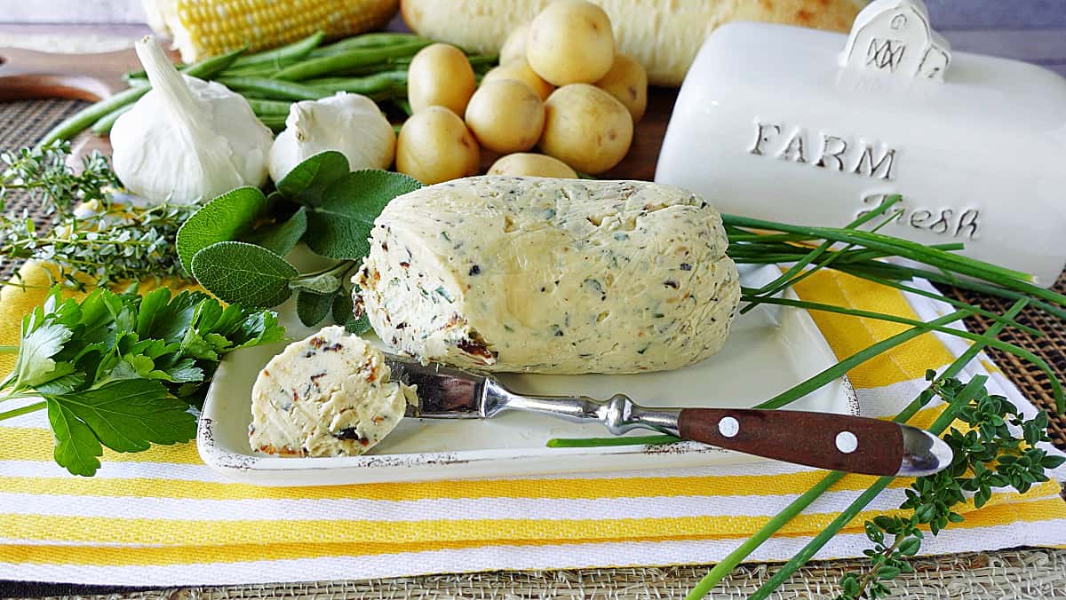 A log of Roasted Garli and Herb Compound butter on a butter dish with a butter knife.