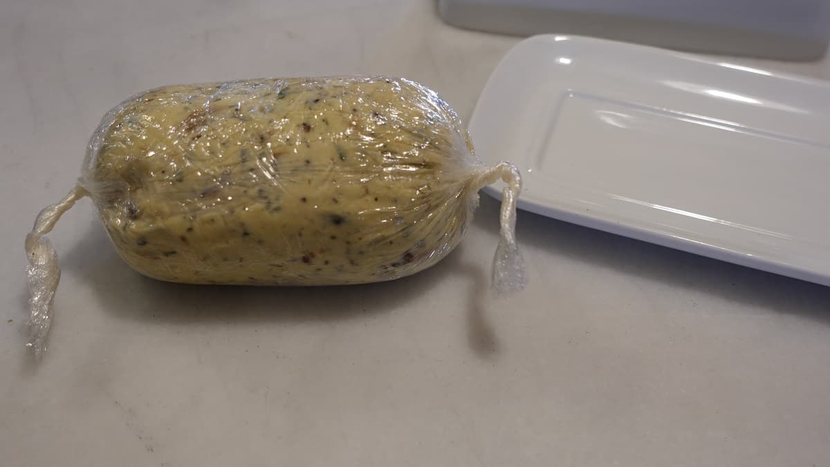 A log of Roasted Garlic Compound Butter with Herbs wrapped in plastic wrap.