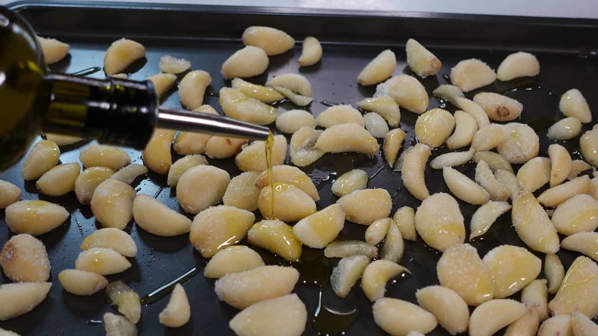 Olive oil being drizzled onto garlic cloves on a baking sheet.