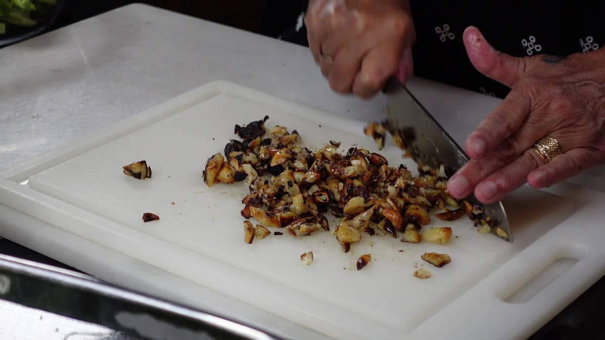 A cutting board with a knife and hands chopping roasted garlic.