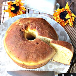 A round loaf of Sally Lunn Batter Bread on a plate with a serrated knife and a slice taken out.