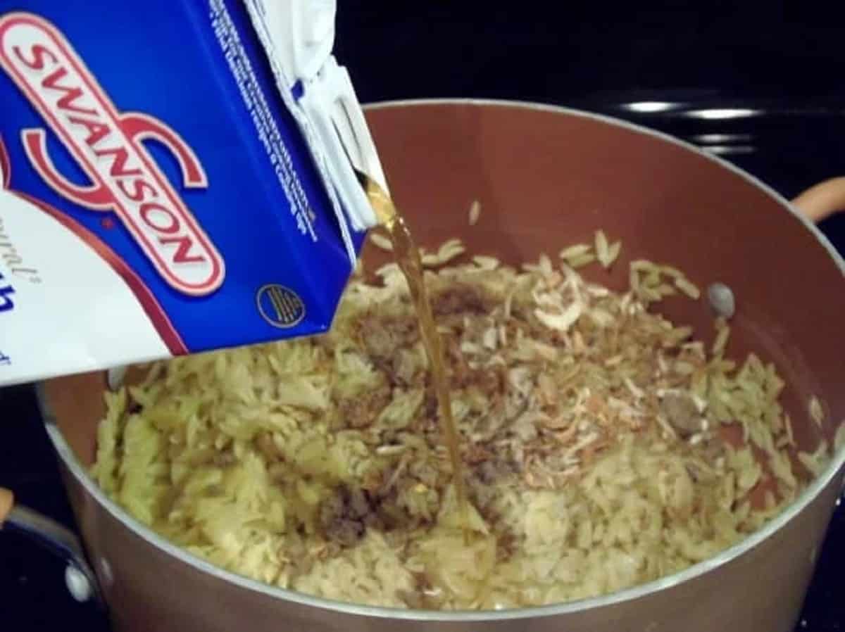 Chicken broth being poured into a saucepan with orzo pasta.