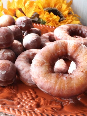 A pile of Glazed Apple Cider Donuts on an orange platter with sunflowers in the background.