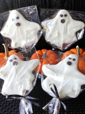 Four White Chocolate Ghost Pops on sucker sticks with mini pumpkins in the background.
