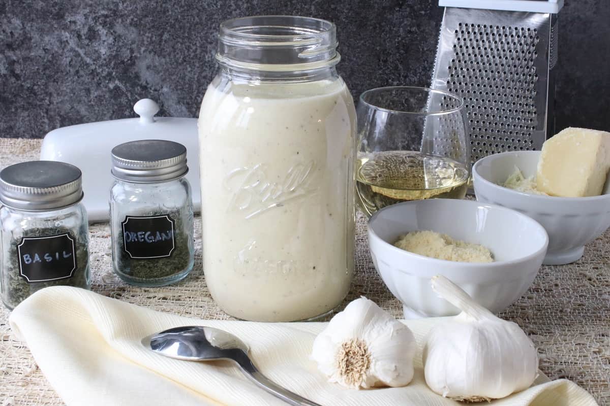 A jar of Homemade White Wine Alfredo Sauce along with a spoon, herbs, garlic and a glass of wine.
