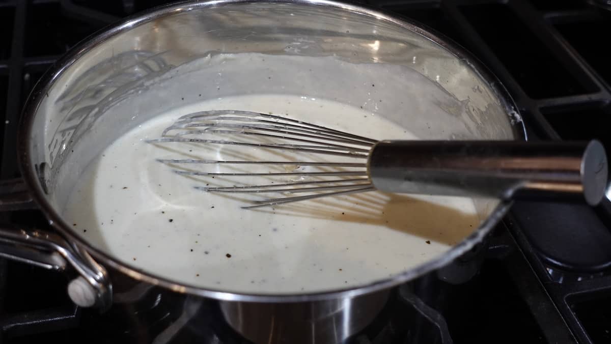 A whisk in a saucepan containing Homemade White Wine Alfredo Sauce.