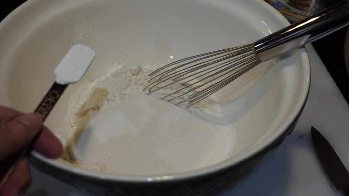 A teaspoon of baking powder being added to a bowl of flour and sugar.