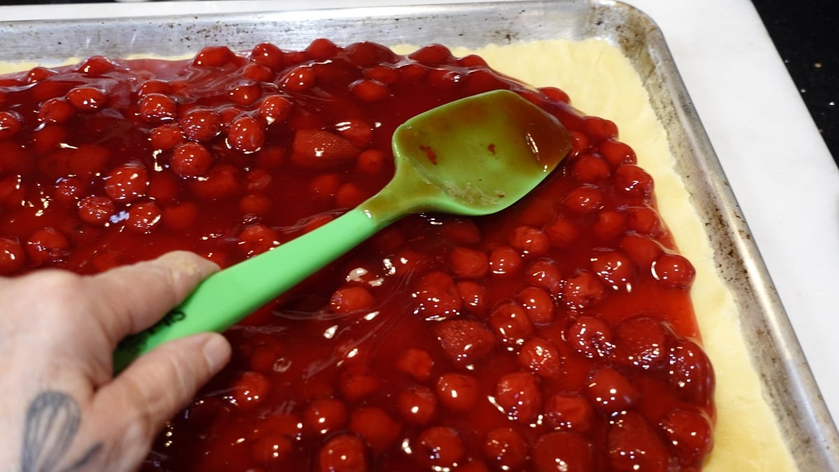 Cherry pie filling being spread over a dough crust with a spatula.