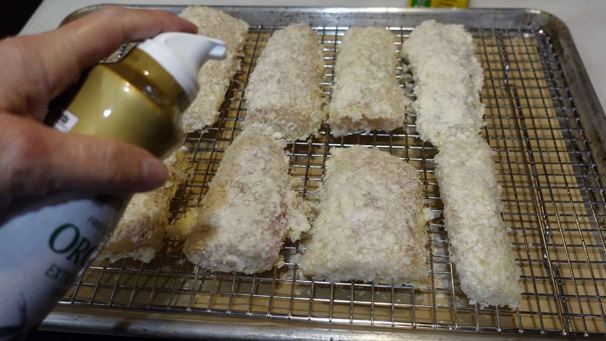 Olive oil spray being applied to Potato Crusted Cod fillets on a baking rack.