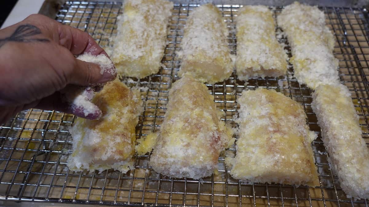 Potato flakes being put onto cod fillets on a baking rack.