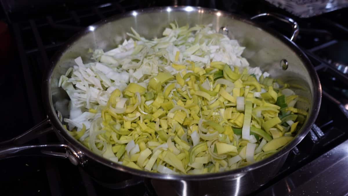 A large skillet on the stovetop filled with shredded cabbage and chopped leeks.