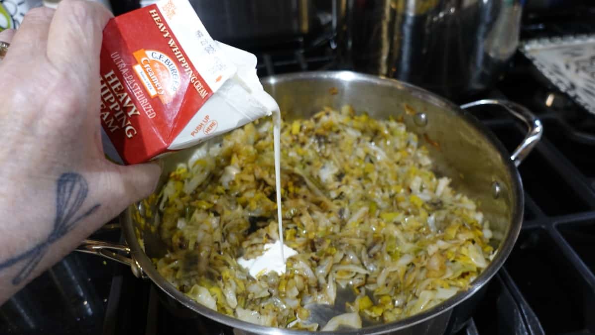 Heavy cream being poured into a skillet with sautéed cabbage and leeks.