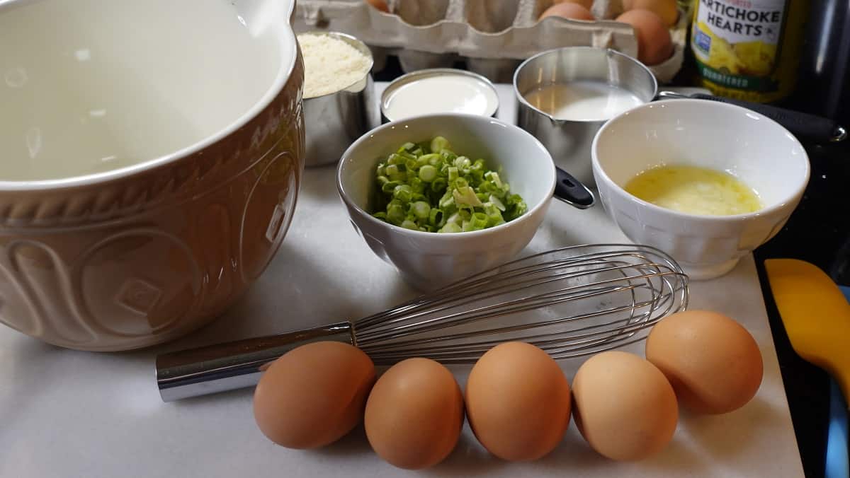 Brown eggs, a whisk, and other ingredients set out to make quiche.