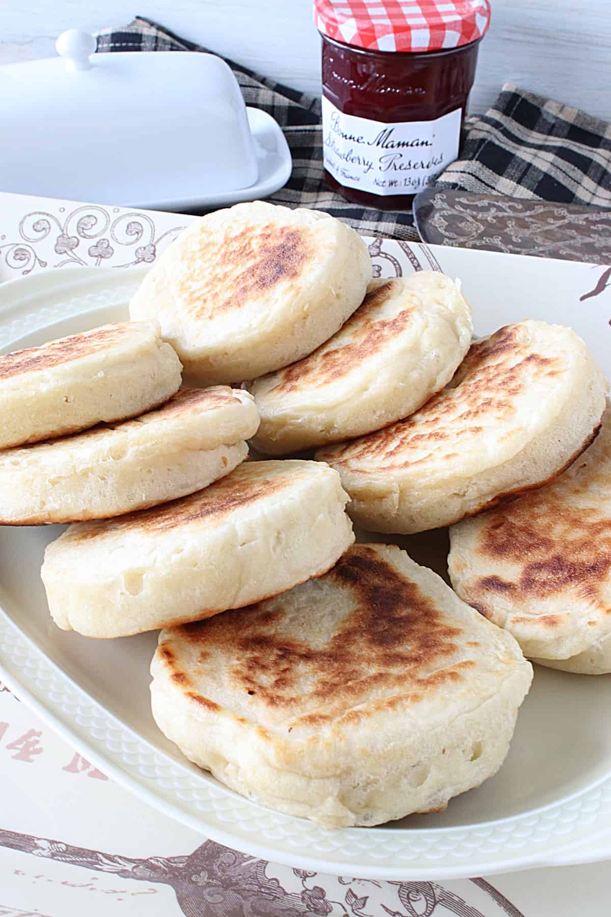 A platter full of Homemade English Crumpets in the forefront with a butter dish and jar of jam in the background.