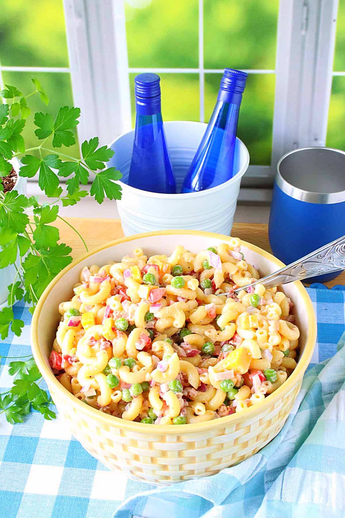A vibrant photo of a bowl of chilled Macaroni and Pea Salad on a blue and white checked tablecloth.