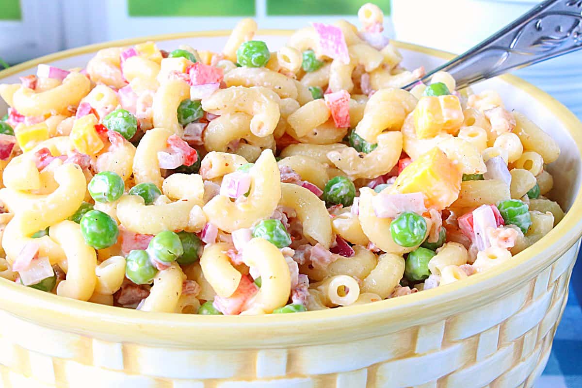 A closeup of a Macaroni and Pea Salad in a ceramic bowl with cheddar cheese cubes and red peppers.