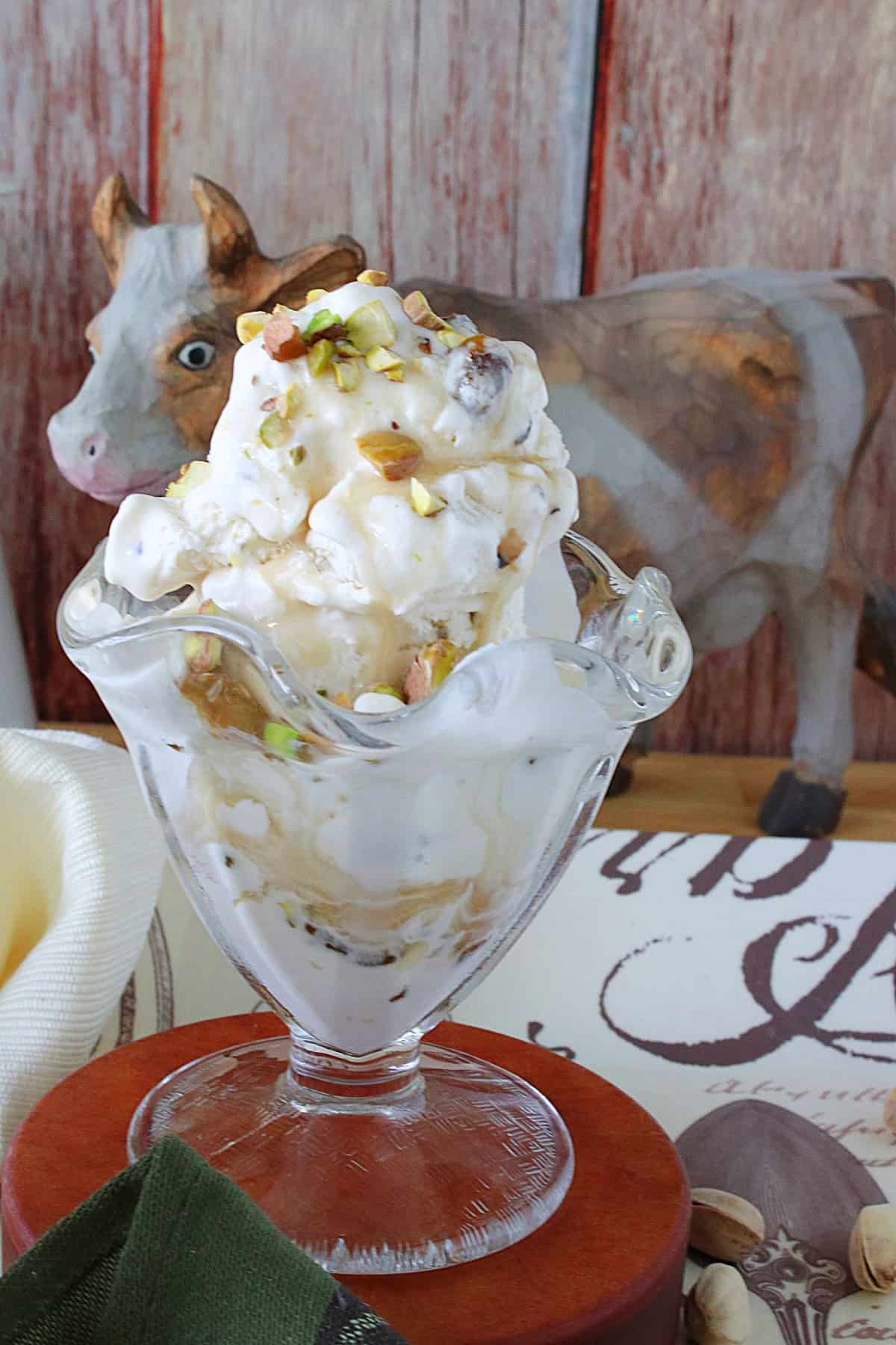 A glass ice cream dish filled with Maple Pistachio Ice Cream in the forefront with a cow in the background.