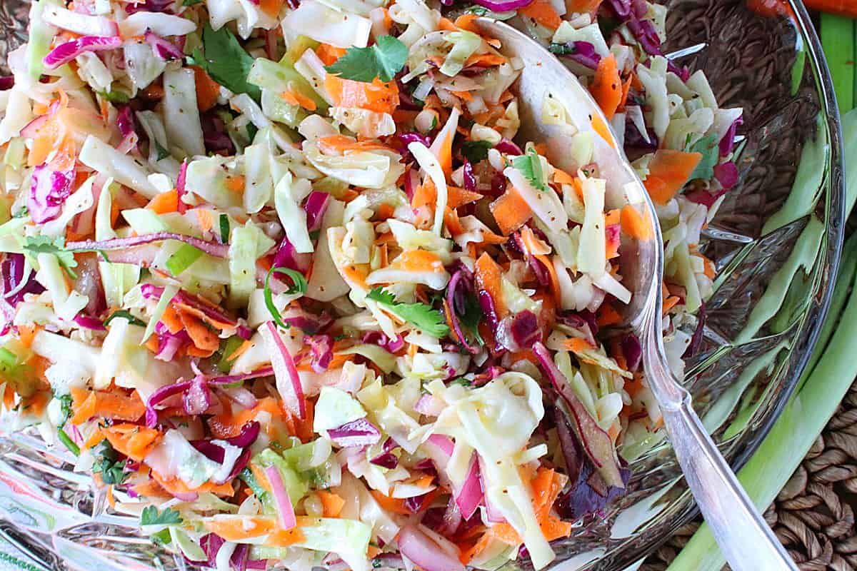 A colorful and crunchy coleslaw in a glass bowl with a silver serving spoon.