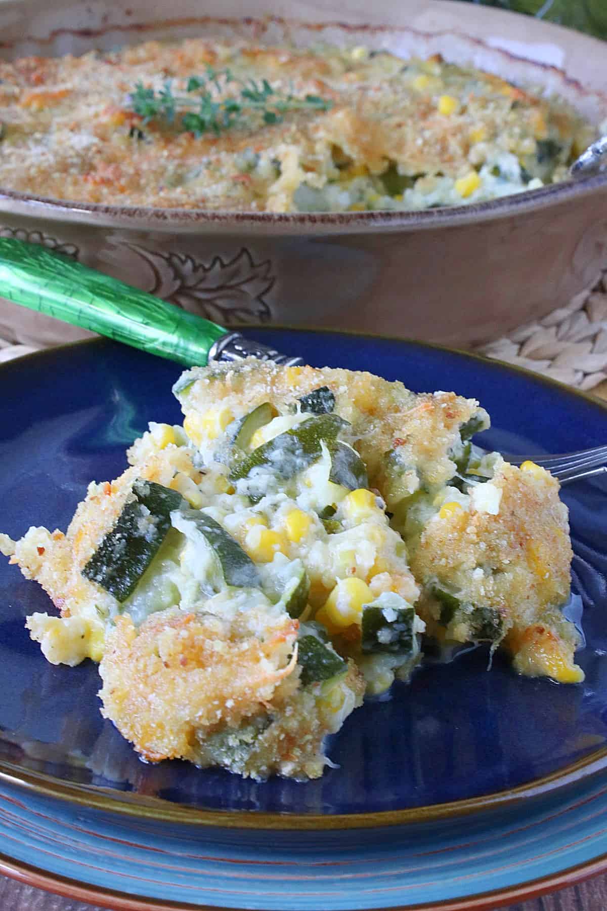 A serving of Zucchini Corn Gratin on a blue plate with a casserole dish in the background. 