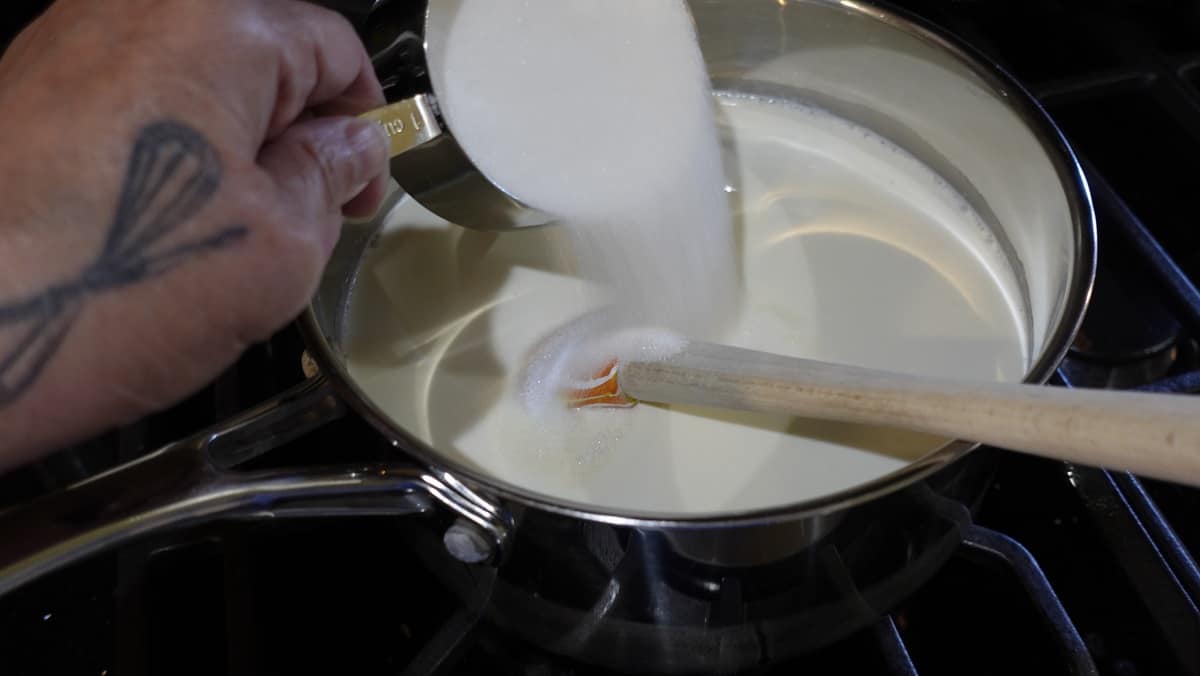 Sugar being added to a saucepan with cream and milk.