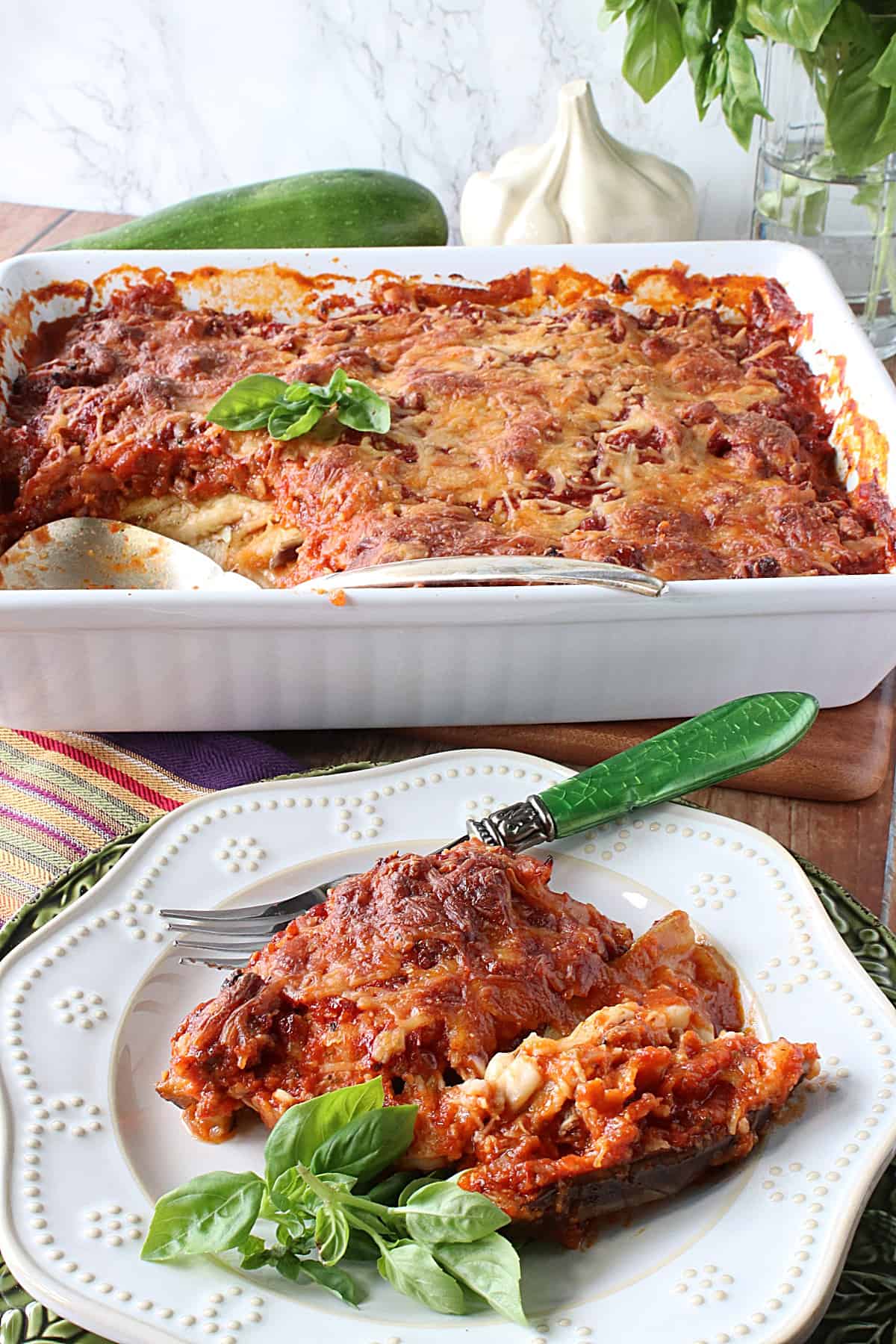 A casserole dish and plate filled with Eggplant and Zucchini Lasagna with sauce and cheese.