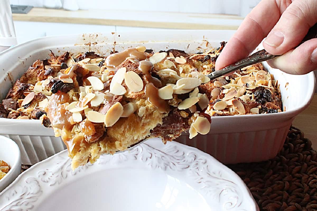 A hand holding a serving spoon with Panettone Bread Pudding covered with almonds and a RumChata caramel sauce. 