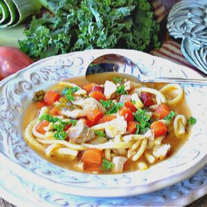 A white bowl filled with a healthy serving of Chicken Noodle Soup with Leeks and Kale along with a spoon.