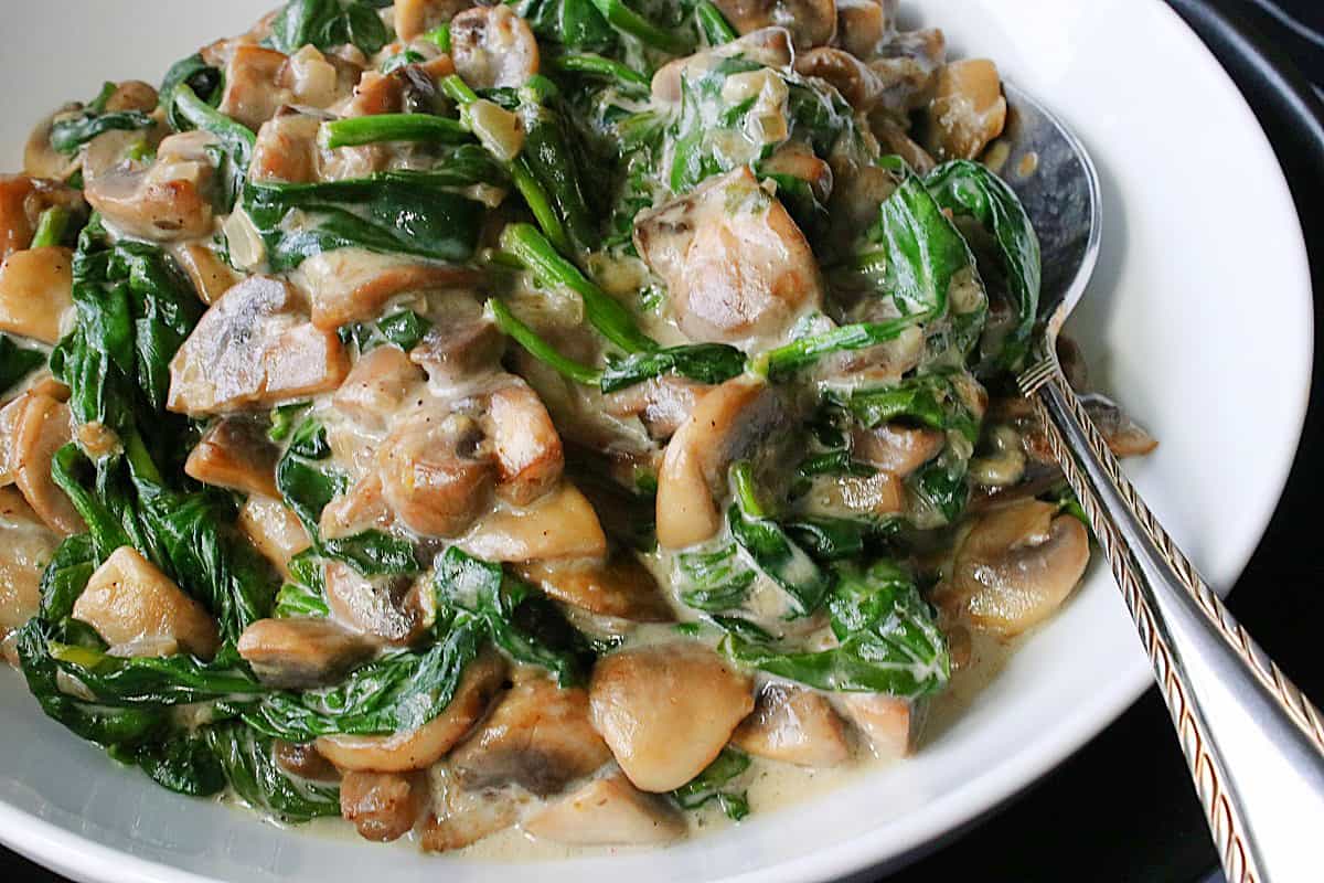 A white serving bowl filled with Creamy Mushrooms and Spinach along with a spoon.