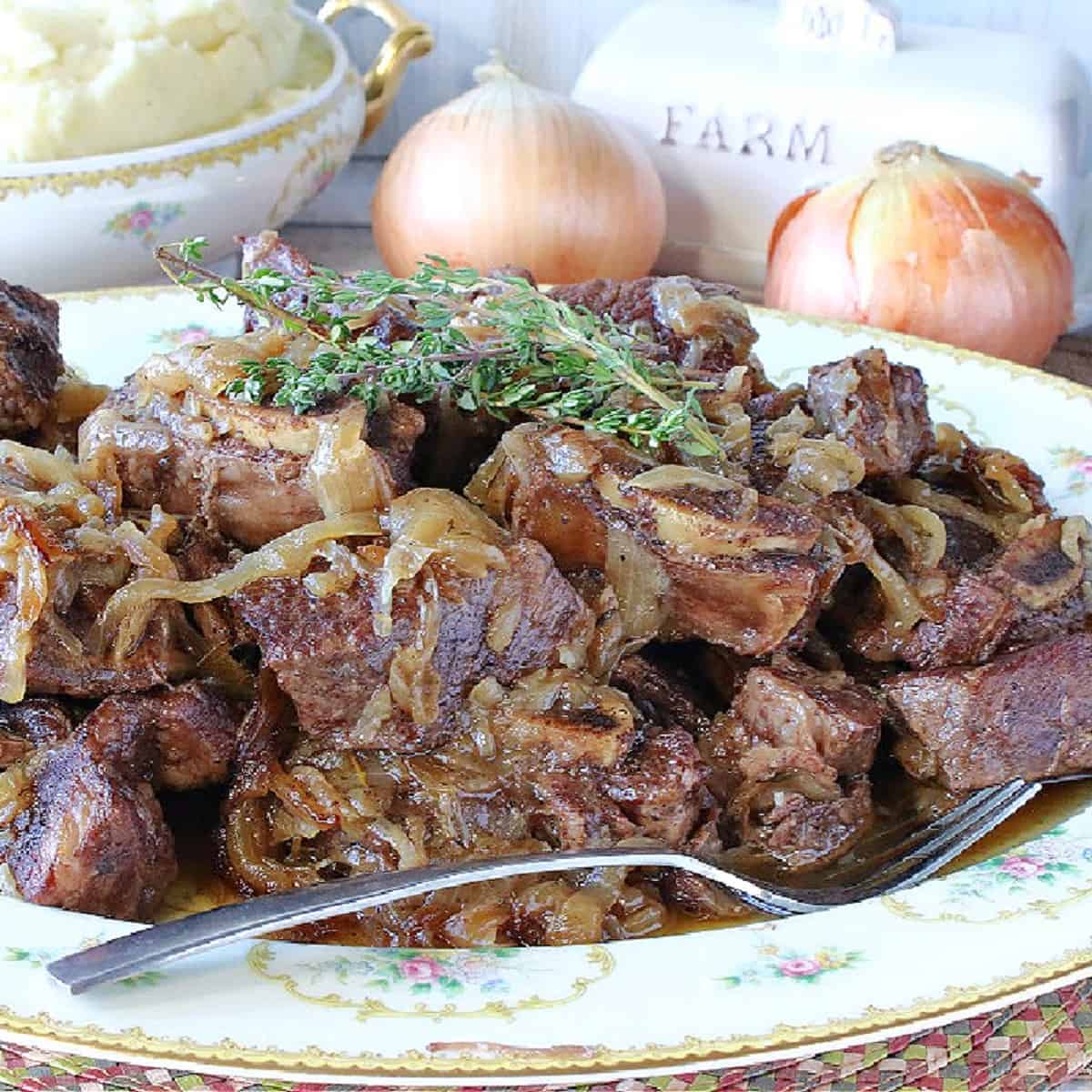 A platter filled with French Onion Short Ribs with fresh thyme sprigs on top and a bowl of mashed potatoes in the background.