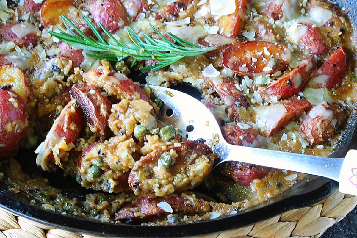 A serving spoon in a cast iron skillet filled with Vesuvio Potatoes and Gravy with fresh rosemary. 