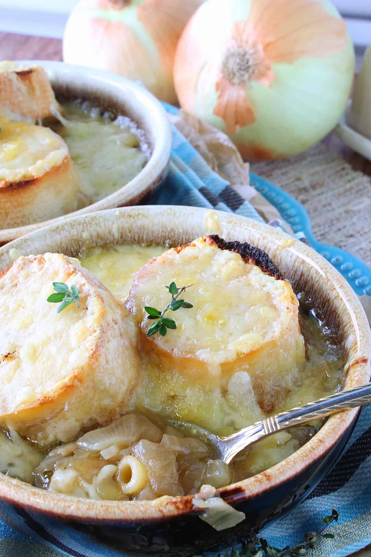 A bowl of French Onion Soup with Pasta along with melted cheese, bread, and a spoon.