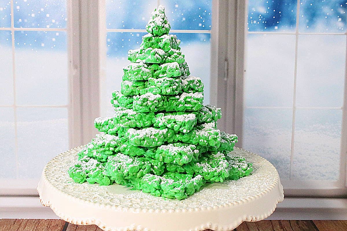 A white cake stand with a 3D Rice Krispies Treats Christmas Tree in front of a snowy window.