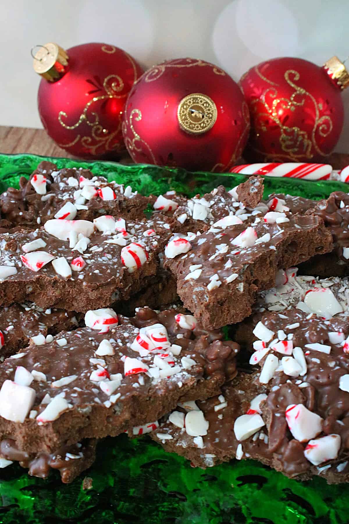 A green glass plate filled with Cocoa Krispies Peppermint Bark with ornaments in the background.