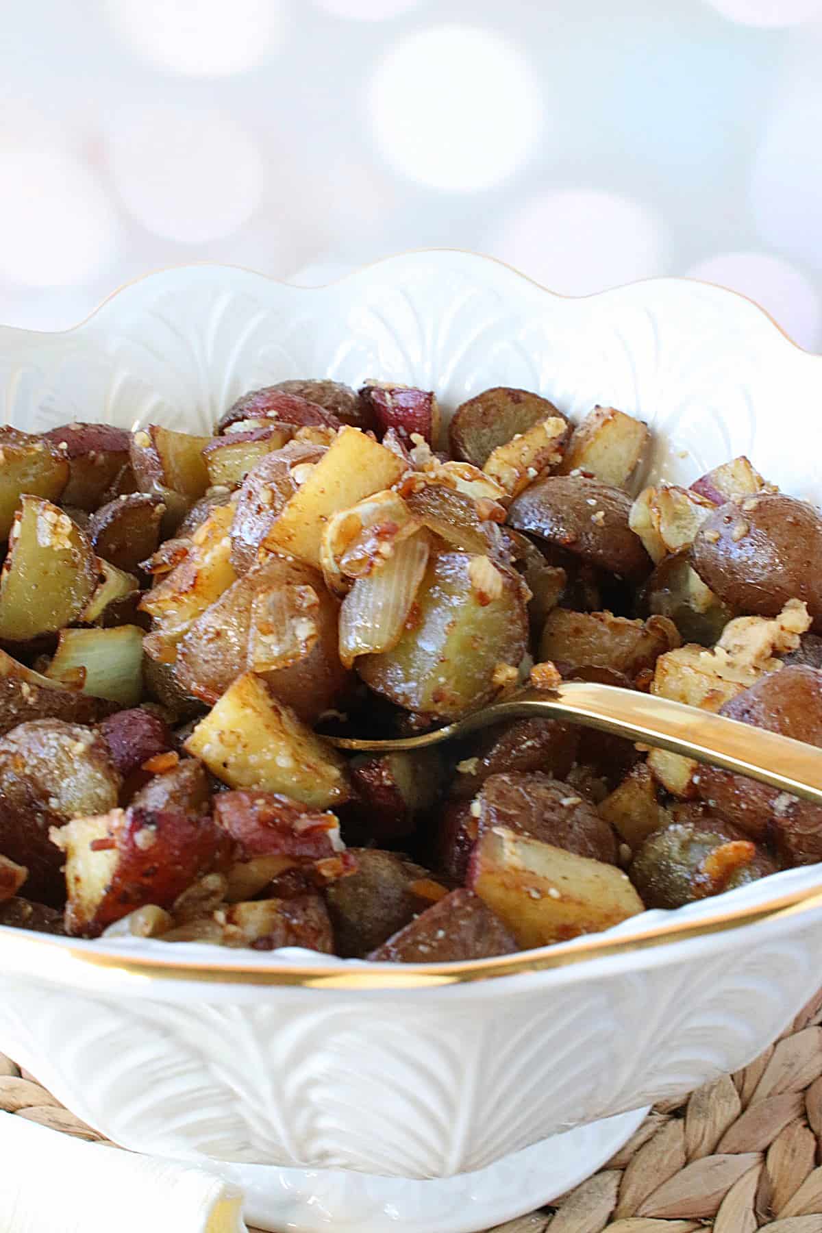 A pretty white serving bowl with a golden spoon filled with Roasted Potatoes with Onion and Parmesan.