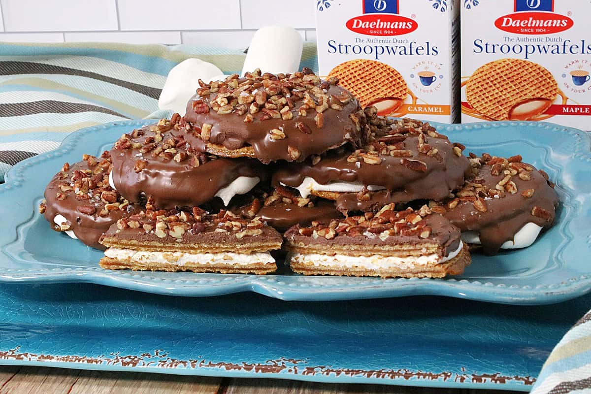 A stack of S'mores Stroopwafel Sandwiches on a blue platter with marshmallows in the background.