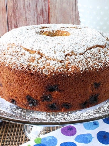 A Sour Cream Poundcake with Blueberry and Orange on a glass cake stand with powdered sugar on top of the cake.