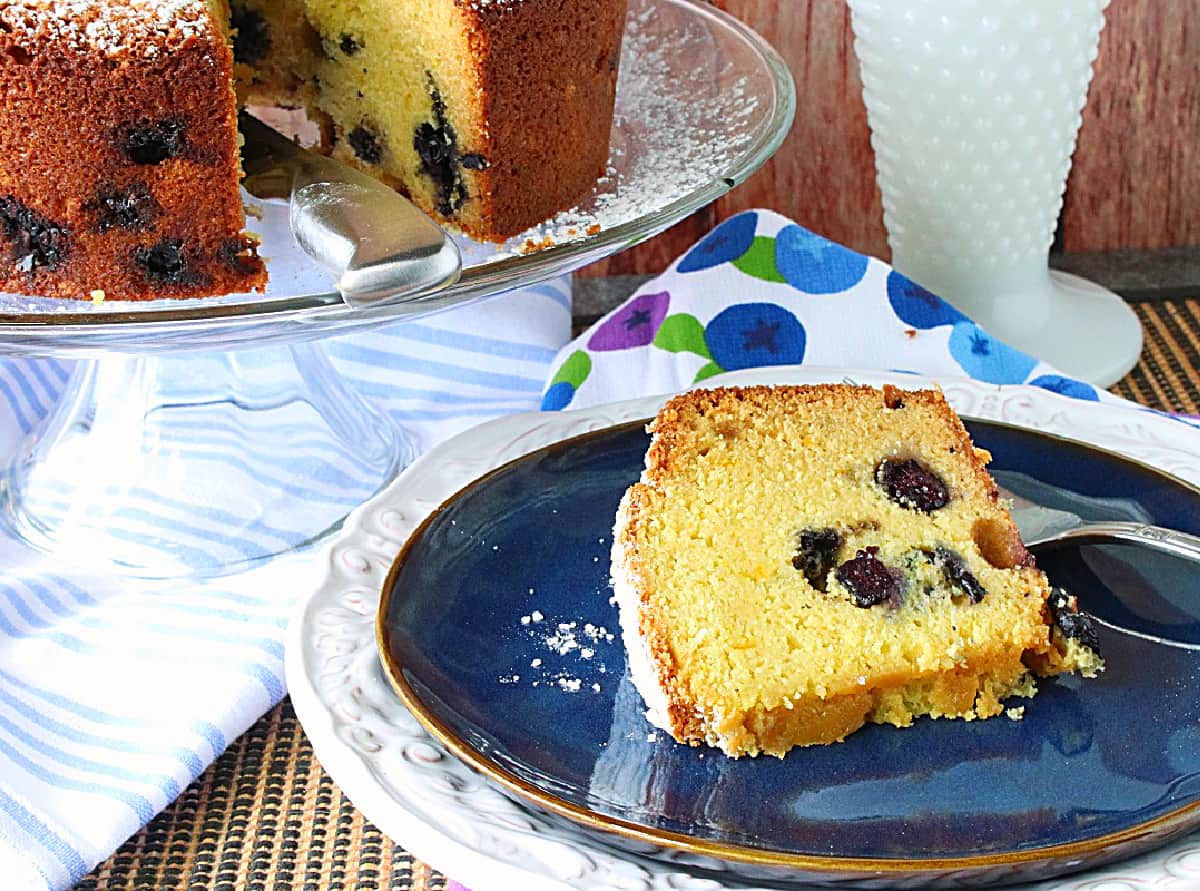 A slice of sour cream poundcake with blueberries and orange on a blue plate with the full cake in the background.
