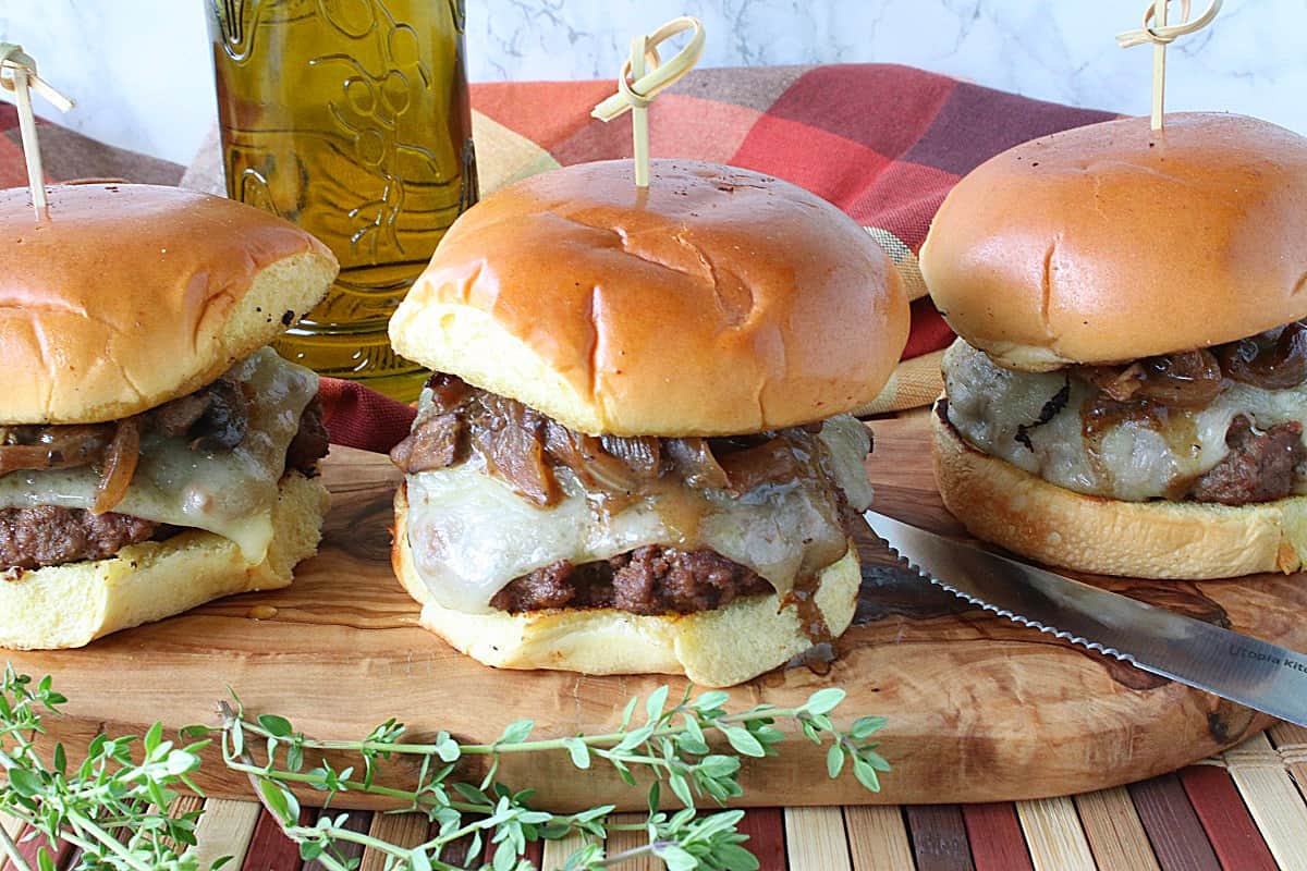 Three Mushroom Swiss Burgers on a wooden board with toothpicks in each burger and a knife on the board.