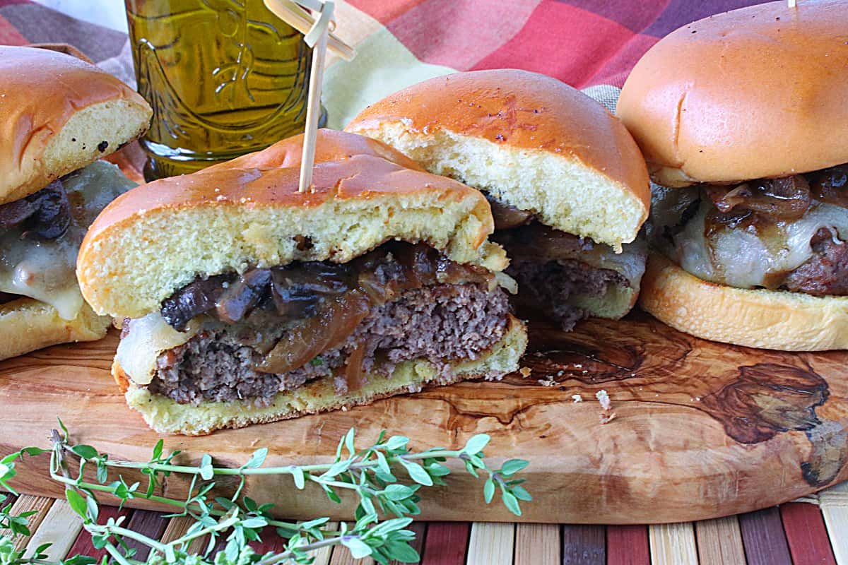 Some Mushroom Swiss Burgers on a wooden board with the front burger cut in half.