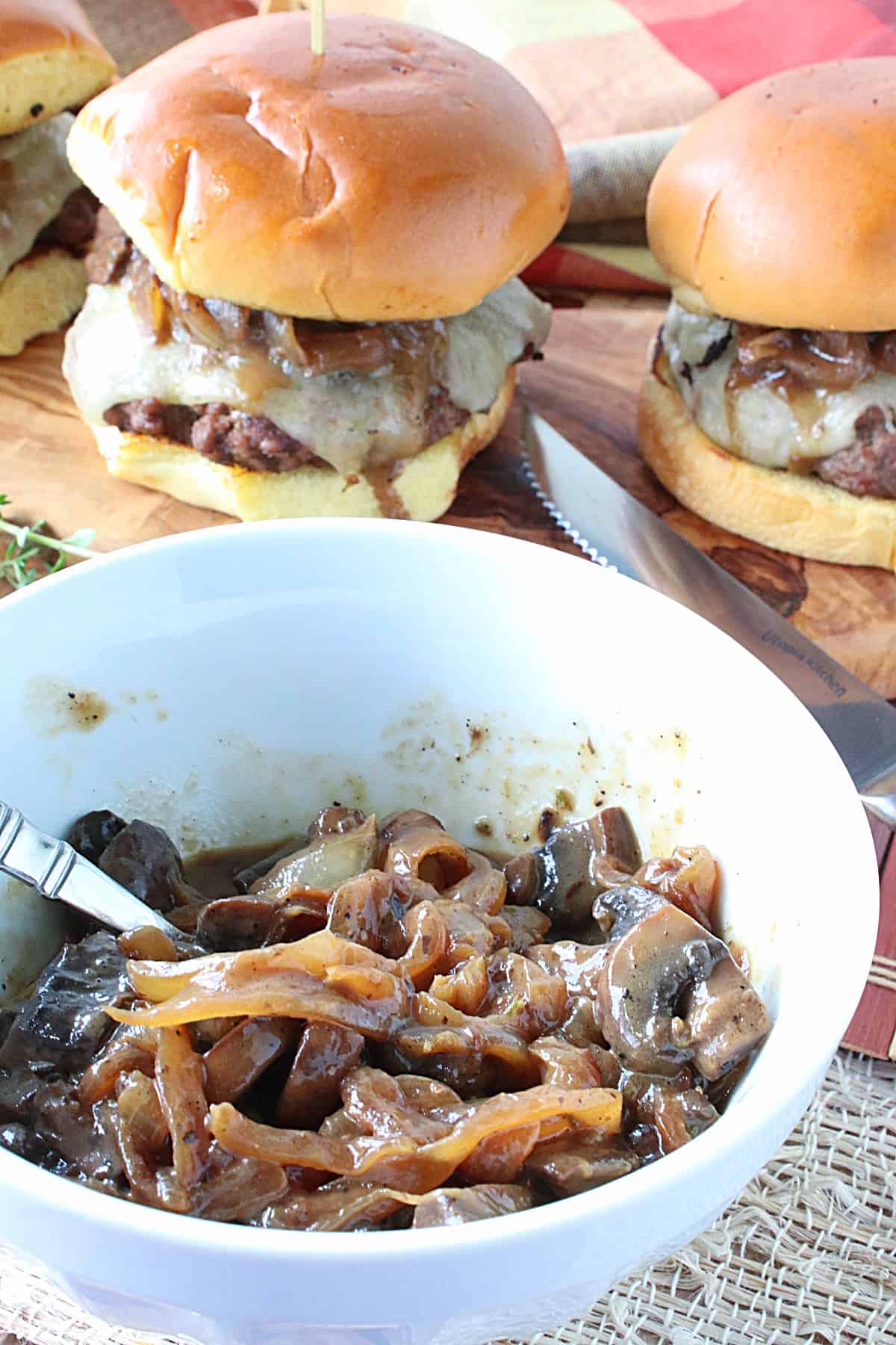 Two Mushroom Swiss Burgers in the background and a white bowl with onions and mushroom topping in the foreground.