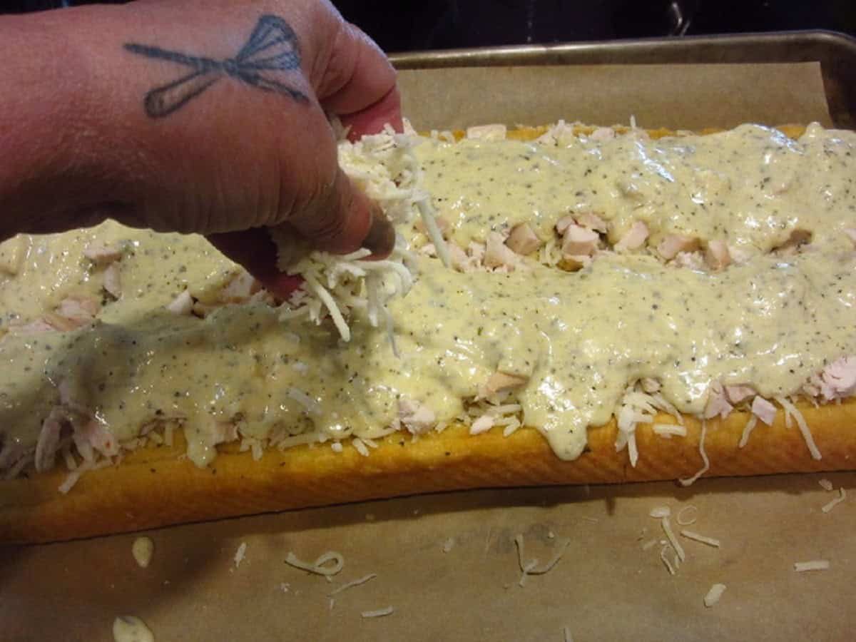 A hand sprinkling shredded mozzarella cheese on top of a Chicken Alfredo Garlic Bread Pizza before baking.