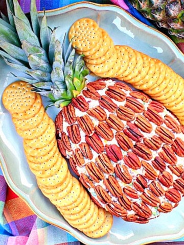 A cute Pineapple Shaped Cheese Ball on a platter surrounded by crackers and a colorful napkin.