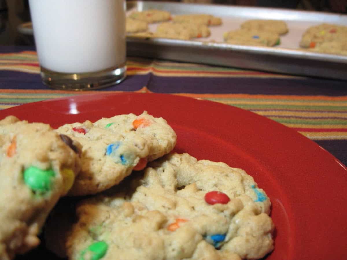A red plate in the foreground and a baking sheet in the background both with mini m and m oatmeal cookies.