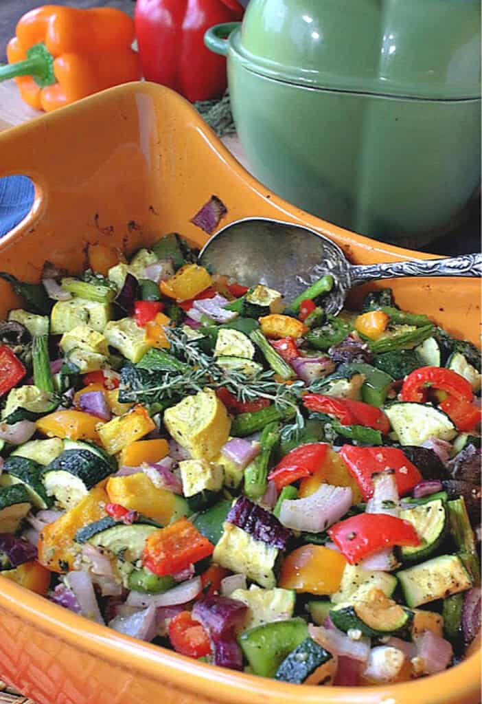 An orange casserole filled with colorful roasted rainbow vegetables with a serving spoon.