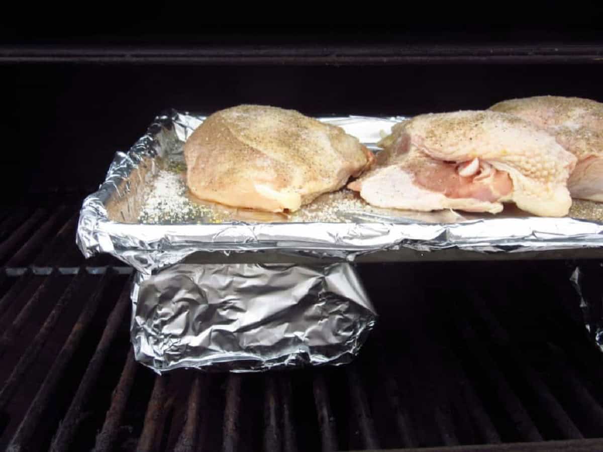 A baking sheet covered with foil and topped with chicken on an outdoor grill with bricks for oven-type cooking.